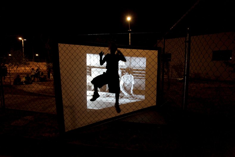 Child climbing a fence is silhouetted by a screen showing a film projected by the Cinecleta, Moviebike, at a park in Saltillo